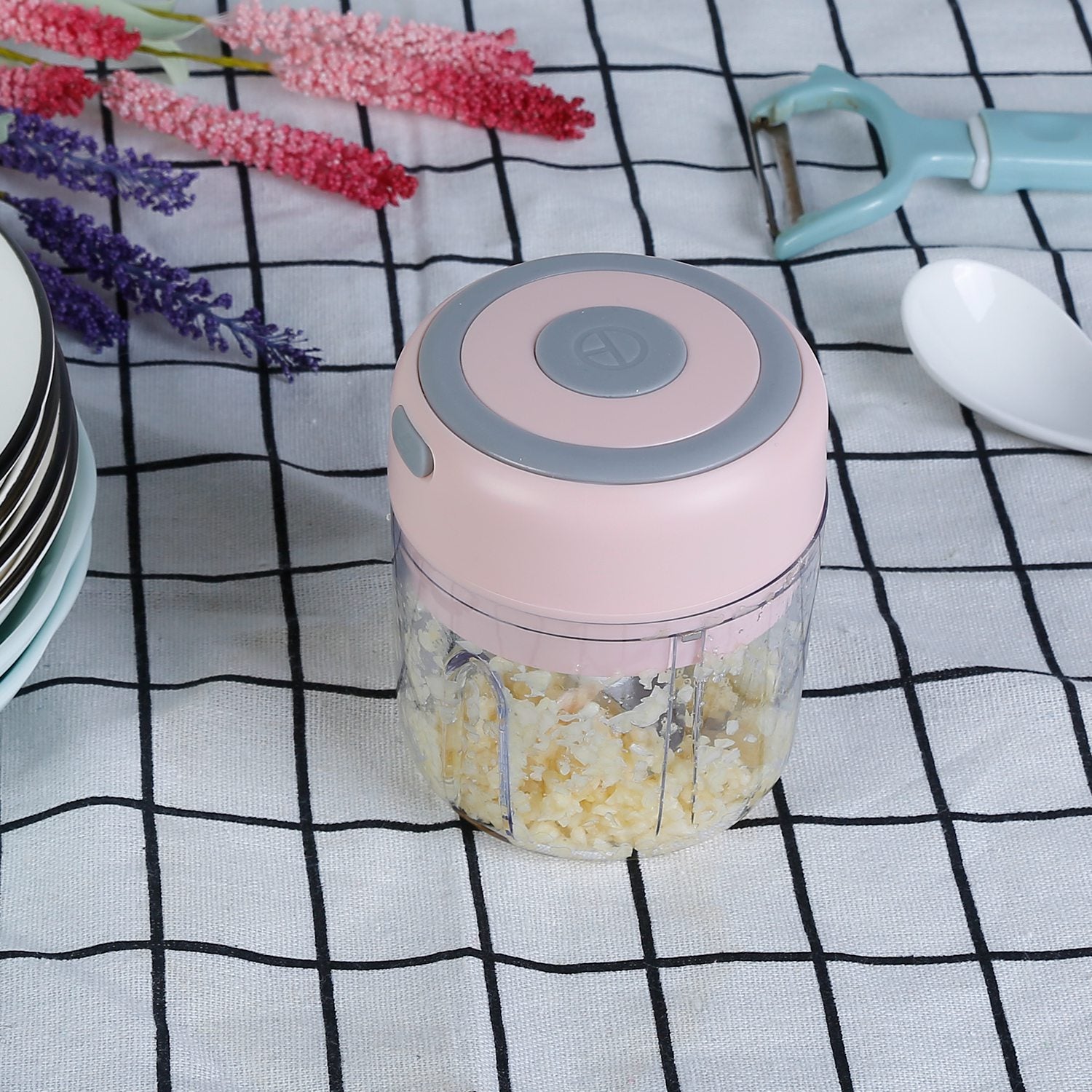 Pink and gray hand blender on a checkered tablecloth with flowers and kitchen tools.